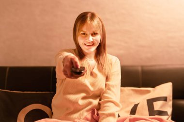 Evdeki kanepede dinlenirken elinde uzaktan kumandayla gülümseyen bir kadın.