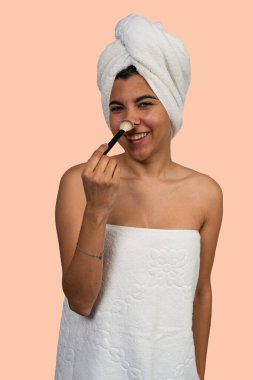 Havlu giymiş, gülümseyen, makyaj fırçasıyla burnuna pudra süren genç bir kadın.