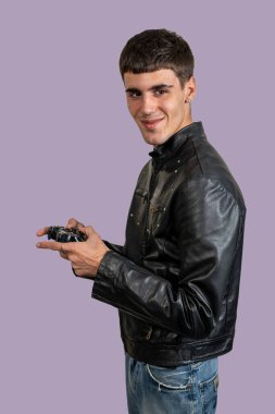 Siyah deri ceket giyen, kablosuz kumanda tutan, video oyunları oynayan, gülümseyen, mor arka planda izole edilmiş bir oyuncu.