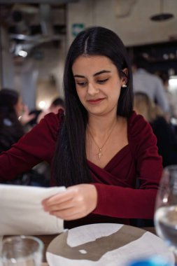 Kırmızı bluzlu genç bir kadın restoranda peçetesini kucağına koyuyor.