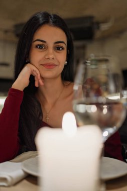 Genç bir kadın gülümsüyor, romantik bir akşam yemeğinin tadını çıkarıyor elinde mum ve bardakla.