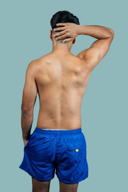 Mavi şortlu gömleksiz genç adam, elleriyle kafasına dokunuyor, arkadan görülüyor.