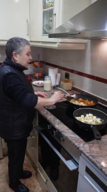 Gri saçlı yaşlı kadın sağlıklı bir yemek hazırlıyor ve tavada sebzeleri karıştırıyor.