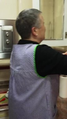 Önlük takan yaşlı bir kadın mutfak dolabını açıyor ve kahve fincanını çıkarıyor.