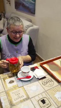 Gülümseyen, gözlüklü, masada oturmuş kendine bir fincan sıcak bitkisel içeceği dolduran son sınıf öğrencisi kadın.