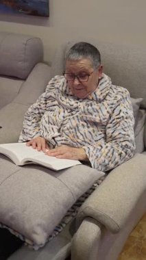 Kısa saçlı ve gözlüklü yaşlı kadın koltukta rahatça otururken kitap okuyor.