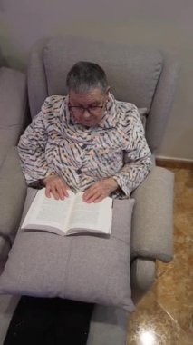 Gözlüklü ve pijamalı yaşlı bir kadın koltukta rahatça oturup evde kitap okuyor.