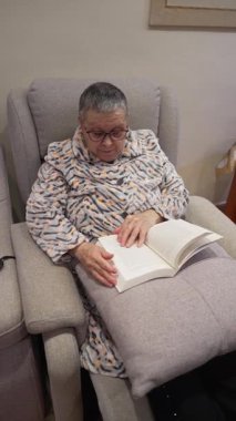 Yaşlı bir kadın gözlük takıyor ve sıcak bir bornoz giyiyor koltukta rahatça oturup kitap okuyor.