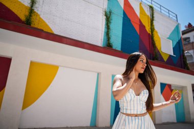 Güneşli bir günde poz veren genç bir kadın, canlı sokak sanatı ve geometrik şekillerle çevrili.