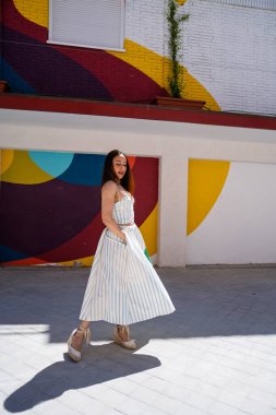 Çizgili iki parçalı yaz kıyafeti giymiş genç bir kadın canlı ve soyut bir duvar resmine karşı poz veriyor.