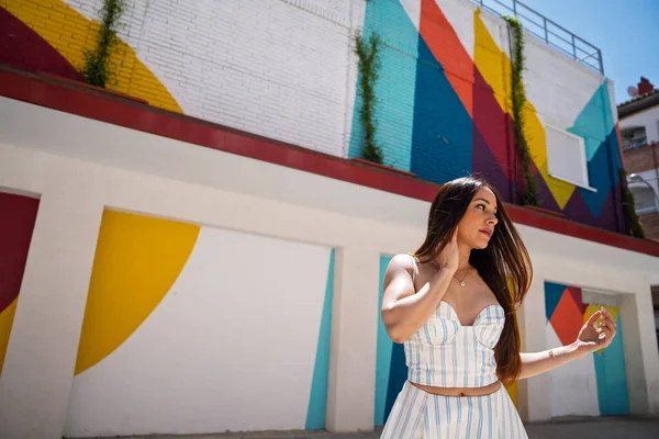 Güneşli bir günde poz veren genç bir kadın, canlı sokak sanatı ve geometrik şekillerle çevrili.