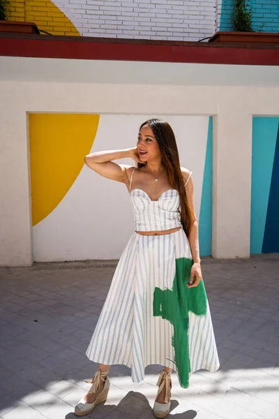 Çizgili iki parçalı yaz kıyafeti içinde renkli bir duvara poz veren genç bir kadın.