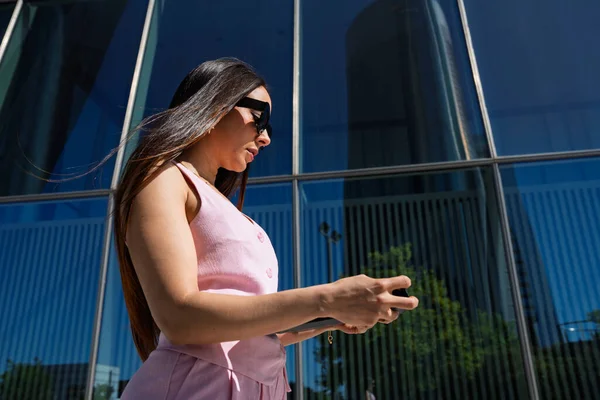 Pembe elbiseli ve güneş gözlüklü bir kadın cam binanın önünde akıllı telefon kullanıyor.