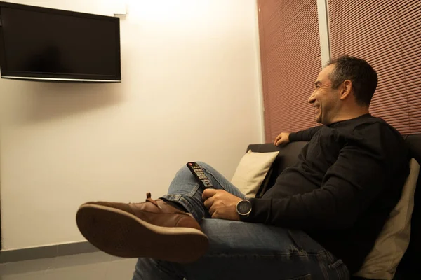 Mutlu adam kanepede uzanıp uzaktan kumandayı tutuyor ve evde televizyon seyrediyor.