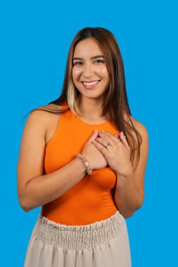 Elleri göğsünde gülümseyerek mavi arka planda şükranlarını ifade eden genç bir kadın.