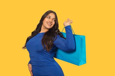 Sarı arka planda mavi bir alışveriş çantası tutan gülümseyen genç kadın.