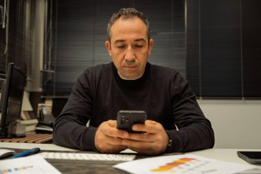 Ofis ortamında bir masada çalışan bir adam cep telefonuna odaklanıyor.