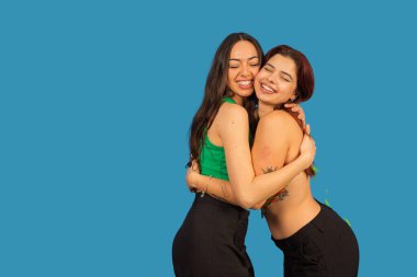 İki gülümseyen kadın mavi arka planda gözleri kapalı birbirlerine sarılıyorlar.