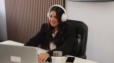 Genç profesyonel bir kadın masasında çalışırken kulaklıkla müzik dinliyor ve gülümsüyor.