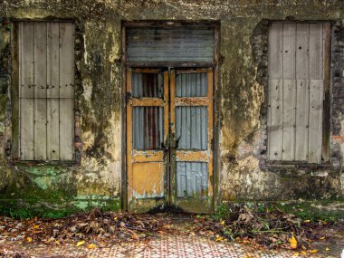 Eski, terk edilmiş, mozaik desenli kiremitleri olan, metal kaplamalı dikdörtgen pencereli bir binanın ön cephesi. Kırık parçaları çinko kaplayan hasarlı ahşap bir kapı. Yıpranmış kırsal duvarlar, kuru yapraklar ve taban..