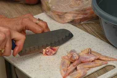 Stüdyoda keten önlüklü bir kadının mutfak masasında sarımsak karanfil, paprika ve zeytinyağı ile kanat keserken çekilen fotoğrafı. Temiz kesikler, pembe et ve sıcak aydınlatma süreci vurguluyor, yemek için mükemmel.