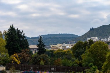 Tepedeki eski bir kilise manzarası ve arkasında dağlar olan Tiflis şehri. Geleneksel mimari ve eski kasaba manzarası yeşil yamaçlar ve dağlık manzarayla harmanlanmış..
