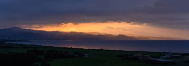 Gün ışığı söndükçe, denizin üzerinde nefes kesici bir renk dizisi belirir, engebeli dağları aydınlatarak manzaraya yumuşak gölgeler bırakır. Huzurlu bir güzellik anı.