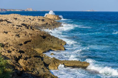 Turkuaz dalgalar, deniz kenarında güneşli bir günde doğanın güzelliğini vurgulayarak kayalık kıyıları kaplar..