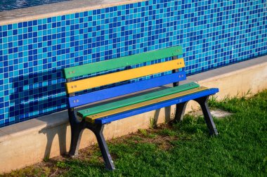 Güneşli bir tatil köyünde havuz kenarına kurulmuş basit bir ahşap bank suyun yanında dinlenmek için huzurlu bir yer sunuyor..