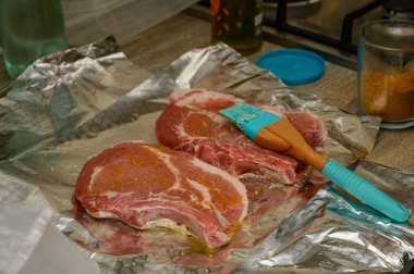 Aromatik baharatla kaplanmış taze domuz pirzolası, pişirmek için hazırlanmış silikon fırçayla dokulu alüminyum folyonun üzerinde dinleniyor. Yakın çekim yemek hazırlama fotoğrafçılığı.