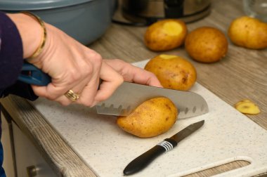 Beyaz kesim tahtasındaki sarı patatesleri dilimlemek için mavi saplı büyük mutfak bıçağı kullanan bir kadının ellerini yakın plan çek. Otantik yemek hazırlama anı.
