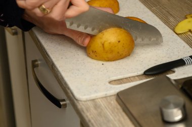 Eller çiğ, derisi yüzülmüş bir patatesi temiz, beyaz bir plastik kesme tahtasıyla büyük, pürüzsüz bir şekilde dilimliyor. Bu resim, ev yapımı haşlanmış patates ve şifalı otlar yapmak için atılan ilk hazırlık adımını gösteriyor..