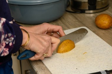 Temiz, beyaz plastik bir kesme tahtasının üzerinde çiğ patatesi kalın ve kırsal dilimleyen ellerin yakın plan görüntüsü. Bu resim ev yapımı patates kızartması ya da fırın kızartması hazırlamak için gerekli ilk adımı gösteriyor..