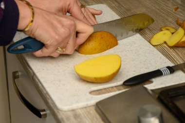Yakın plan çekimde, çiğ, soyulmamış bir patatesi açık renkli plastik bir kesme tahtasının üzerinde kalın, kırsal bir kama haline getiriyorlar. Patates kabuğunun dokusuna ve basit, pratik mutfak görevine odaklan..