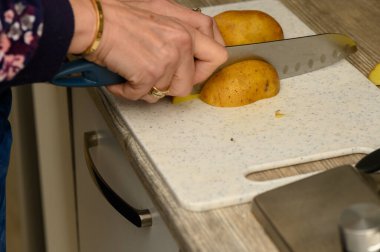 Beyaz plastik bir mutfak tahtasında çiğ patatesi büyük ve pürüzlü dilimlere ayıran ellerin yakın çekimi. Bu görüntü, ev yapımı kavrulmuş patates yemekleri yapmak için elle hazırlama aşamasını yakalar.