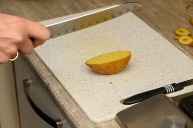 Beyaz plastik bir kesme tahtasının üzerinde çiğ, soyulmamış bir patatesi kalın takozlara ayıran ellerin yukarıdaki görüntüsü. Patates kabuğunun doğal dokusuna ve klasik bir meze hazırlama sürecine odaklanın.