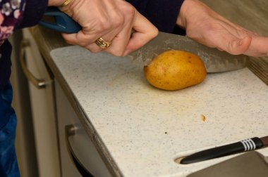 Yakın plan çekimde, açık renkli plastik bir kesme tahtasının üzerinde çiğ, soyulmamış bir patatesi kalın takozlara ayırıyorlar. Odak noktası, patatesin doğal şekli ve elle hazırlanmış yiyecek hazırlama süreci..