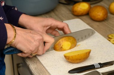Beyaz plastik bir kesme tahtasının üzerinde kirli bir patatesi kalın takozlara ayıran ellerin yakın çekimi. Bu resim, ev yapımı kızarmış patates veya patates yapmak için temel bir mutfak görevini yakalar..