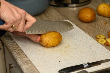 Beyaz plastik bir kesme tahtasının üzerinde çiğ patatesi düz olmayan bir şekilde dilimleyen el fotoğrafları. Bu resim, kırsal patates yemekleri için temel yemek hazırlama adımını gösteriyor..