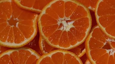 Yukardan aşağı, bir daire şeklinde dizilmiş taze mandalina dilimlerinin makro çekimi, karanlık bir levha ya da desenli bir yüzey üzerinde yavaşça dönüyor. Görüntüler parlak, sulu bölümleri vurguluyor. Canlı turuncu renkler.