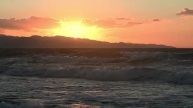 Kıbrıs manzarası üzerinde canlı bir günbatımını yakalayan nefes kesici 4K hava görüntüsü. Çekim, dağların dramatik siluetlerini (Troodos aralığı) turuncu, pembe bir gökyüzüne karşı çerçeveler.
