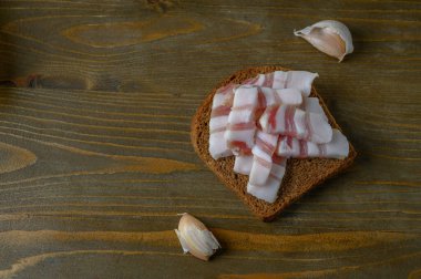 Üzerinde bir dilim kurutulmuş salo sosu olan koyu çavdar ekmeğinin minimalist kompozisyonu kaba, rustik ahşap bir tahtanın üzerinde dinleniyor. Çiğ dokuları, toprak tonlarını ve geleneksel basitliği vurgular.