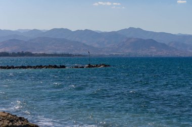 Dalgalar yavaşça kayalık kıyılara vururken dağlar ihtişamlı bir şekilde arka planda parlak mavi bir gökyüzünün altında yükselirken.
