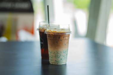 Plastik bardaklarda iki buzlu içecek, ön planda katmanlı bir kibrit latte bulunan karanlık bir masaya konur..