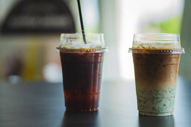 Plastik bardaklarda iki buzlu içecek, ön planda katmanlı bir kibrit latte bulunan karanlık bir masaya konur..