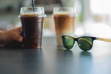 El ele tutuşun. Güneş gözlüğü ve plastik bardaklarda iki buzlu içecek koyu bir masanın üzerine yerleştirilmiş, ön planda katmanlı bir matcha latte yer alıyor..
