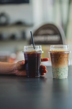 Bir el, koyu bir masada, katmanlı, buzlu bir kibritli lattenin yanında bir fincan buzlu siyah kahve tutar..