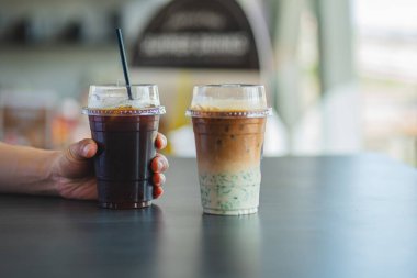 Bir el, koyu bir masada, katmanlı, buzlu bir kibritli lattenin yanında bir fincan buzlu siyah kahve tutar..