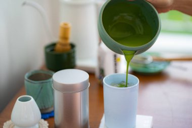 Yeşil kibrit çayı, çay yapma aletleriyle çevrili beyaz bir fincana dökülüyor..