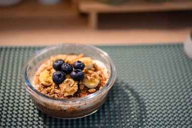 Temiz bir cam kasede güzel katmanlı kahvaltı parfümü, kremalı yoğurt, gevrek granola, taze muz dilimleri, ve desenli deniz mavisi silikonlu bir paspasın üzerine konmuş dolgun yaban mersinli bir taç..